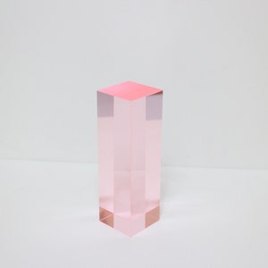 Könnte beinhalten: Ein klarer rosa Acrylblock, ein rechteckiger Prisma, mit einer glatten Oberfläche. Der Block steht auf einer weißen Oberfläche.