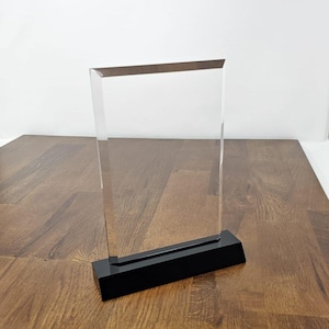 May include: A clear acrylic award plaque with a black base. The plaque is rectangular and has a beveled edge. The base is made of a solid black material.