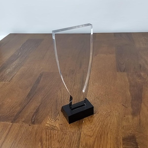 Könnte beinhalten: Eine klare Acrylauszeichnung mit schwarzem Sockel. Die Auszeichnung hat die Form eines Schildes mit einem abgerundeten oberen und unteren Rand.