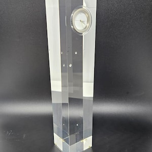 May include: A clear acrylic clock with a white face and black hands. The clock is standing on a black surface.