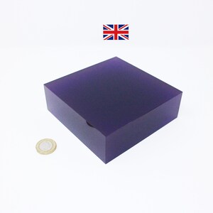 Könnte beinhalten: Ein quadratischer Block aus durchsichtigem, violettem Acrylharz. Der Block ist etwa so groß wie eine 1-Euro-Münze.