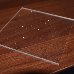 May include: A clear sheet of acrylic with a sparkly, glittery finish. The sheet is lying on a brown wooden surface.