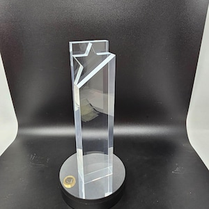 Könnte beinhalten: Eine transparente Acryl-Auszeichnung mit einem sternförmigen Design oben. Die Auszeichnung ist rechteckig und steht auf einem schwarzen, kreisförmigen Sockel. Eine Münze ist auf dem Sockel sichtbar. Der Hintergrund ist schwarz.