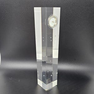 May include: A clear acrylic clock with a white face and black hands. The clock is encased in a rectangular block of clear acrylic with small air bubbles throughout.