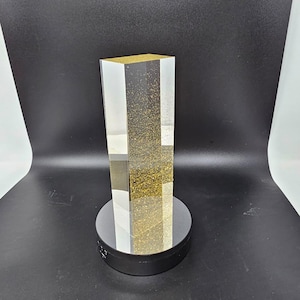 May include: A clear, rectangular acrylic block with gold glitter suspended inside, mounted on a black circular base. The top of the block is also filled with gold glitter. The object is a decorative item.