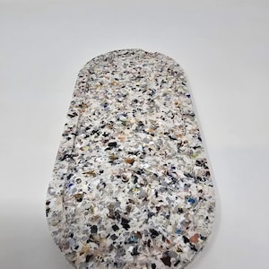 May include: A white oval-shaped coaster made from recycled materials. The coaster is covered in small, colourful pieces of recycled materials, creating a speckled pattern.