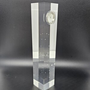 May include: A clear acrylic clock with a white face and black hands. The clock is encased in a rectangular block of acrylic with small air bubbles throughout.