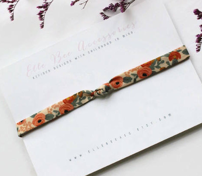 May include: A white card with the text "Elle Bee Accessories" and "Refined Designs with Childhood in Mind." A floral patterned fabric hair tie is tied around the card. The text "www.ellebee.etsy.com" is printed on the bottom of the card.