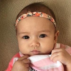 May include: A baby girl wearing a pink and white striped shirt with a floral headband. The headband has a bow in the front.