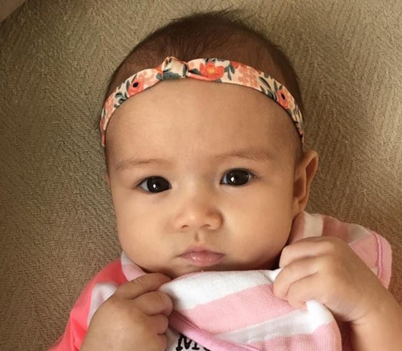 May include: A baby girl wearing a pink and white striped shirt with a floral headband. The headband has a bow in the front.