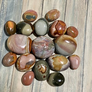 May include: A selection of polished stones in diverse shapes and sizes. The stones exhibit a spectrum of colours, including brown, pink, grey, and green, with distinctive patterns and swirls. The stones are arranged on a light wood-grain surface.