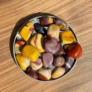 May include: A collection of polished, multi-colored stones in a silver bowl. The stones are a mix of red, yellow, brown, and purple.