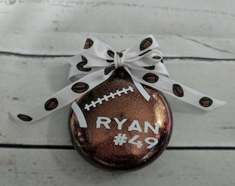 May include: A football-shaped ornament with a glittery brown finish. The ornament features the name "RYAN" and the number "49" in white. A white ribbon with football designs is tied in a bow at the top, adding a decorative touch.