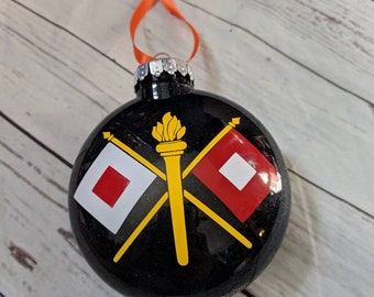 May include: Black round Christmas ornament with a silver cap and orange ribbon. The ornament features a yellow torch and two crossed flags, one with a red square and the other with a white square.