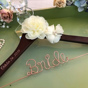 以下が含まれることがあります： 「Bride」という文字がローズゴールドのワイヤーで形作られ、小さなハートがあしらわれたダークブラウンの木製ハンガー。ハンガーには「October 26, 2019」という日付が刻印されており、クリーム色の布製の花が2つ飾られています。