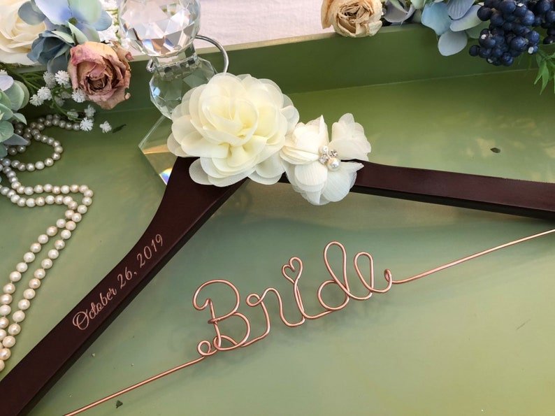 以下が含まれることがあります： 「Bride」という文字がローズゴールドのワイヤーで形作られ、小さなハートがあしらわれたダークブラウンの木製ハンガー。ハンガーには「October 26, 2019」という日付が刻印されており、クリーム色の布製の花が2つ飾られています。