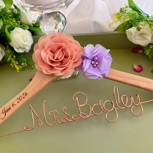 以下が含まれることがあります： Mrs. Bagleyと書かれた銅線のスクリプトと、June 6, 2026という日付が刻印された木製ハンガー。ハンガーには、大きなピーチ色の布製フラワーと、中央にクリスタルのアクセントが付いた小さなラベンダー色の布製フラワーが飾られています。