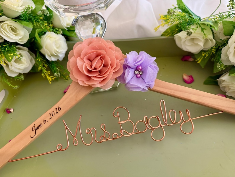 以下が含まれることがあります： Mrs. Bagleyと書かれた銅線のスクリプトと、June 6, 2026という日付が刻印された木製ハンガー。ハンガーには、大きなピーチ色の布製フラワーと、中央にクリスタルのアクセントが付いた小さなラベンダー色の布製フラワーが飾られています。