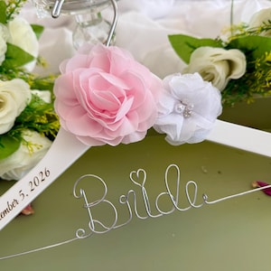 以下が含まれることがあります： Brideという文字を形作った銀色のワイヤーと小さなハートがあしらわれた白い木製ハンガー。ハンガーには大きなピンクの布製バラと小さな白い布製花が飾られています。2026年9月5日という日付が印字されています。