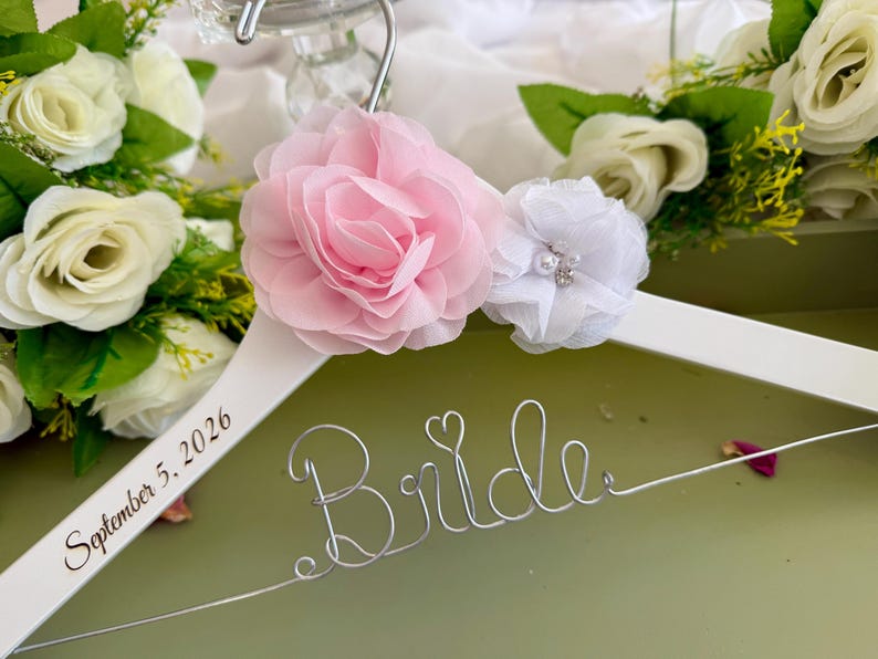 以下が含まれることがあります： Brideという文字を形作った銀色のワイヤーと小さなハートがあしらわれた白い木製ハンガー。ハンガーには大きなピンクの布製バラと小さな白い布製花が飾られています。2026年9月5日という日付が印字されています。