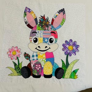 Könnte beinhalten: Ein farbenfrohes Patchwork-Esel-Applikation mit großen Ohren, einem lächelnden Gesicht und zwei Blumen. Der Esel besteht aus verschiedenen gemusterten Stoffen in Rosa-, Blau-, Gelb- und Grüntönen. Die Blumen sind rosa und lila mit grünen Stielen und Blättern.