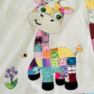 May include: A colorful patchwork giraffe applique on a white background. The giraffe features a patchwork body with various floral and geometric patterns in shades of pink, green, yellow, and blue. A bee and a flower are also appliqued on the white background.
