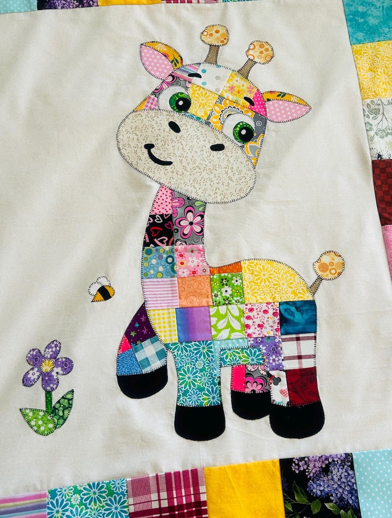 May include: A colorful patchwork giraffe applique on a white background. The giraffe features a patchwork body with various floral and geometric patterns in shades of pink, green, yellow, and blue. A bee and a flower are also appliqued on the white background.