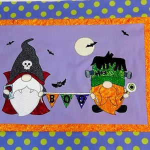 May include: A Halloween-themed fabric panel featuring two gnome characters. One is dressed as a vampire, the other as Frankenstein. The word "BOO" is displayed on a banner between them. The panel has a purple background with a moon and bats.