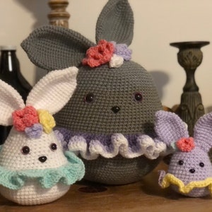 May include: Three crocheted bunny plushies with different colored fur and floral accents. The largest bunny is gray, the middle bunny is white, and the smallest bunny is purple.