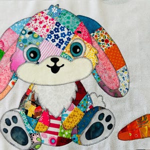 May include: A patchwork fabric applique of a rabbit with long ears, constructed from various colorful patterned textiles including floral, polka dot, and striped prints. The rabbit has a cream-colored face and belly, and a separate orange carrot applique.