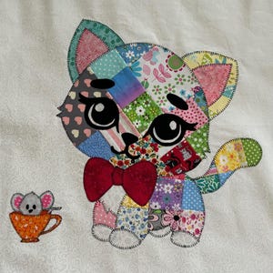 Op de afbeelding: Een kleurrijke patchwork kattenapplicatie met een rode strik en grote zwarte ogen. Een kleine muis in een oranje theekopje staat naast de kat. De kat is gemaakt van verschillende stoffen met patronen, waaronder bloemen- en hartmotieven, gestikt op een witte achtergrond.