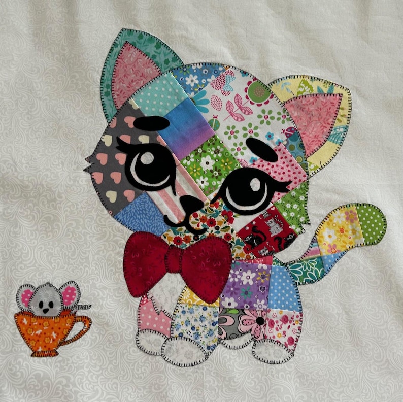 Op de afbeelding: Een kleurrijke patchwork kattenapplicatie met een rode strik en grote zwarte ogen. Een kleine muis in een oranje theekopje staat naast de kat. De kat is gemaakt van verschillende stoffen met patronen, waaronder bloemen- en hartmotieven, gestikt op een witte achtergrond.