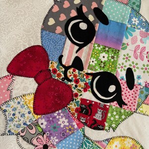Op de afbeelding: Een kleurrijk patchwork-ontwerp met een cartoon kattengezicht met grote zwarte ogen en een rode strik. Het gezicht van de kat is samengesteld uit verschillende stoffen vierkanten met bloemen-, hart- en streepjespatronen. De randen zijn omrand met zwarte stiksels.