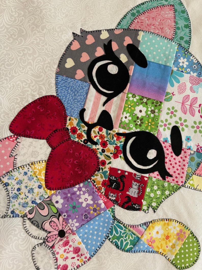 Op de afbeelding: Een kleurrijk patchwork-ontwerp met een cartoon kattengezicht met grote zwarte ogen en een rode strik. Het gezicht van de kat is samengesteld uit verschillende stoffen vierkanten met bloemen-, hart- en streepjespatronen. De randen zijn omrand met zwarte stiksels.
