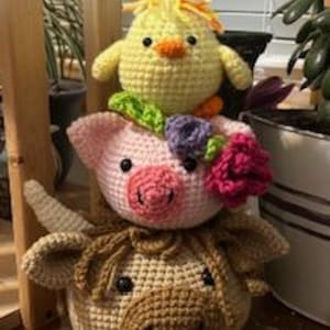 Puede incluir: Una pila de tres animales de peluche tejidos a crochet: una vaca marrón, un cerdo rosa con flores moradas y un pollito amarillo.