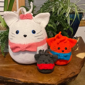 May include: Three crocheted cat plushies. The largest plush is white with pink accents and blue eyes. The middle plush is gray with red accents. The smallest plush is orange with blue accents.