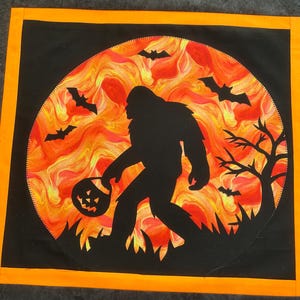 May include: A Halloween-themed square quilt featuring a black silhouette of Bigfoot carrying a jack-o'-lantern against a fiery orange and yellow background. Bats and a bare tree are also silhouetted. The quilt has a black center with an orange border.