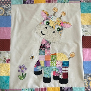 May include: A colorful patchwork giraffe applique on a white background, surrounded by a patchwork border. The giraffe has a smiling face, green eyes, and is accompanied by a bee and a flower. The quilt is a charming handmade item.