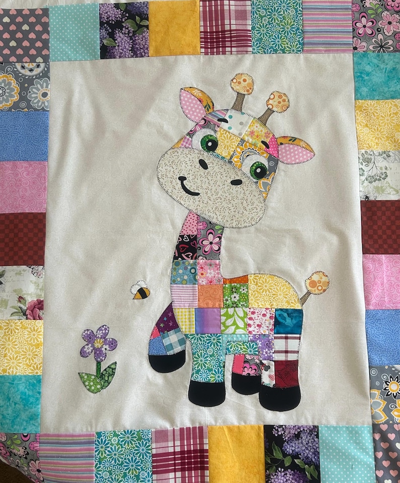 May include: A colorful patchwork giraffe applique on a white background, surrounded by a patchwork border. The giraffe has a smiling face, green eyes, and is accompanied by a bee and a flower. The quilt is a charming handmade item.