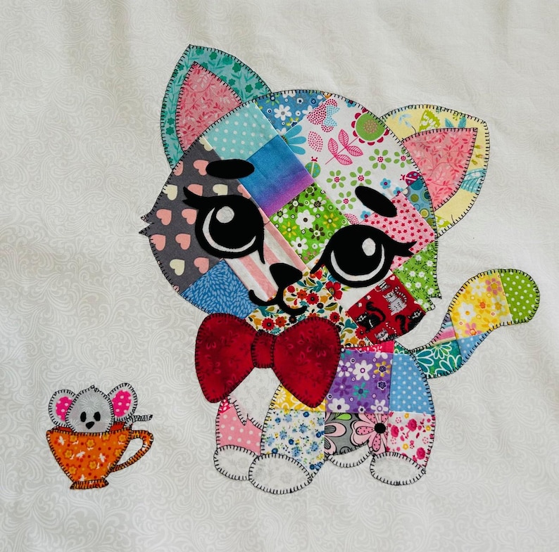 Op de afbeelding: Een kleurrijke patchwork kat met een rode strik en grote zwarte ogen. De kat is gemaakt van verschillende stoffen met patronen. Een kleine muis in een oranje theekopje staat naast de kat. De achtergrond is wit met een subtiel patroon.