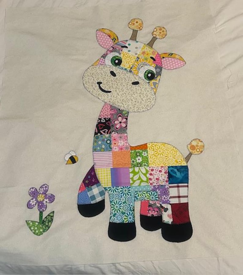 May include: A colorful patchwork giraffe applique with a smiling face, green eyes, and small ears. A bee and a flower are next to the giraffe. The giraffe is made of various patterned fabrics, including floral and geometric designs.