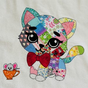 Op de afbeelding: Een kleurrijke patchwork kat met een rode vlinderdas en grote zwarte ogen. De kat is gemaakt van verschillende stoffen met patronen, waaronder bloemen- en stippenontwerpen. Een kleine muis in een oranje theekopje staat naast de kat.