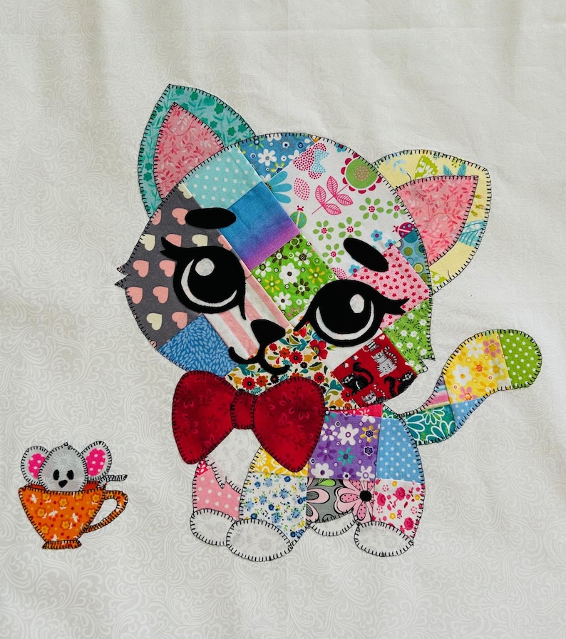 Op de afbeelding: Een kleurrijke patchwork kat met een rode vlinderdas en grote zwarte ogen. De kat is gemaakt van verschillende stoffen met patronen, waaronder bloemen- en stippenontwerpen. Een kleine muis in een oranje theekopje staat naast de kat.