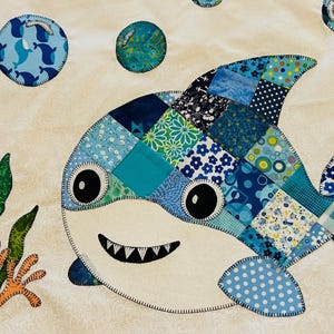 May include: A colourful patchwork shark design in blues, teals, and white. The shark has a patchwork body, large eyes, and a smiling mouth. Bubbles and seaweed accents complete the underwater scene. The design is on a white background.