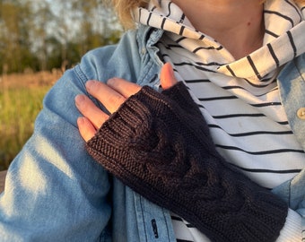 May include: Black fingerless gloves with a cable knit pattern. The gloves are worn on the hand of a person wearing a denim jacket and a striped shirt.