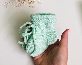 May include: A single, pale green knitted baby bootie with a lace-up design. The bootie is held in a hand, highlighting the soft texture and delicate craftsmanship. The background is a clean white surface with dried flowers.