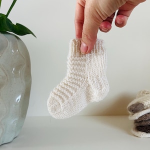 Op de afbeelding: Een kleine, crèmekleurige gebreide babysok die door een hand wordt vastgehouden. De sok heeft een geribbelde boord en een textuurpatroon op de voet. Een stapel soortgelijke sokken is zichtbaar op de achtergrond. Thema: babykleding.