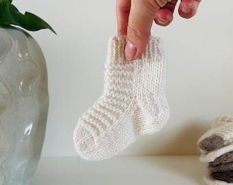 May include: A small, off-white knitted baby sock held by a hand. The sock has a ribbed cuff and textured pattern on the foot. A stack of similar socks is visible in the background. The theme is baby clothing.