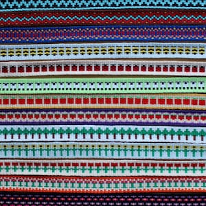 Pode incluir: Uma coleção de fitas tecidas coloridas com vários padrões, incluindo formas geométricas, flores e árvores. As fitas estão dispostas em filas sobre uma superfície marrom clara.