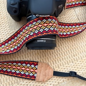 Pode incluir: Uma câmera Canon EOS 700D preta com uma alça estampada. A alça apresenta um design geométrico em vermelho, laranja, amarelo e branco. A alça da câmera é feita de tecido tecido e tem extremidades de couro.